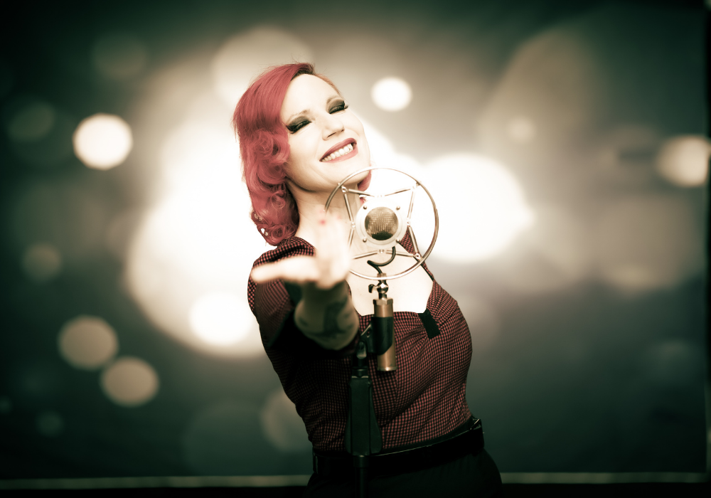 A person with red hair sings passionately into a microphone. The background features glowing bokeh lights, creating a vibrant and energetic atmosphere.