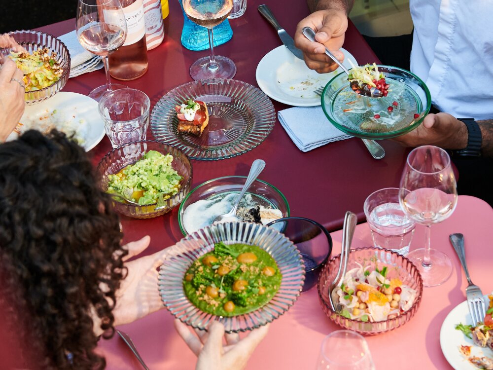 A vibrant table scene with diverse dishes on colorful glass plates, including salads, ripe avocado, and pomegranate seeds. People share and enjoy food.