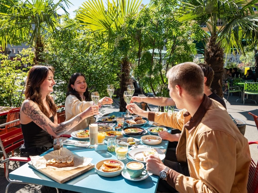 Four people toast with glasses of sparkling wine at an outdoor brunch. The table is filled with colorful dishes and drinks, surrounded by lush greenery.