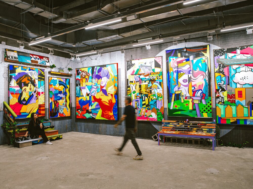 A vibrant art gallery with colorful pop art paintings on the walls. A person sits on steps, another walks by, conveying a lively, urban ambiance.