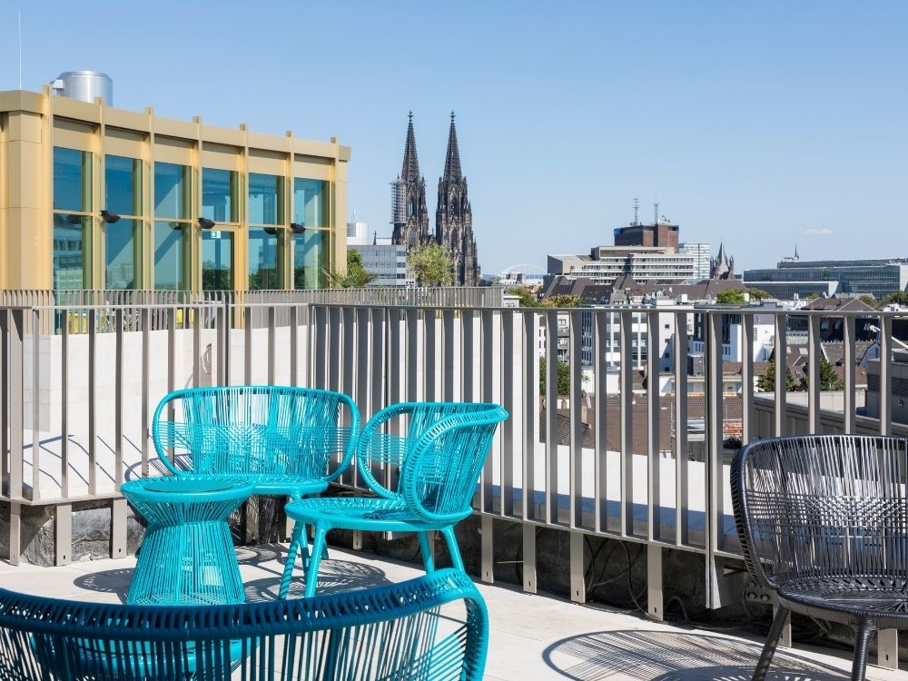 A rooftop terrace with blue woven chairs and tables overlooks a cityscape, including a distant view of the twin spires of Cologne Cathedral.