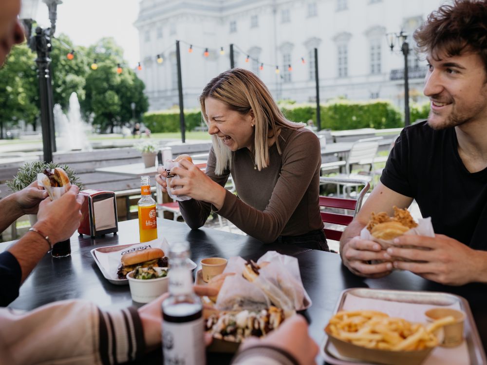 Three people sitting outdoors at a table, enjoying drinks and burgers, with a city street and buildings in the background.