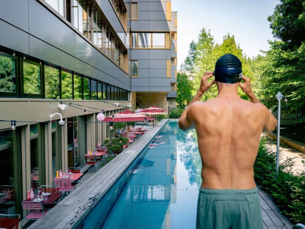 A man prepares to swim in a sleek, narrow pool beside a modern building. Red parasols line the side, with lush greenery and blue sky visible.