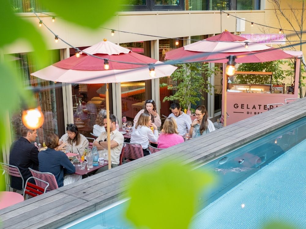 Outdoor restaurant scene with people gathered around tables under red umbrellas, enjoying drinks. Festive string lights and a gelato stand create a lively atmosphere.