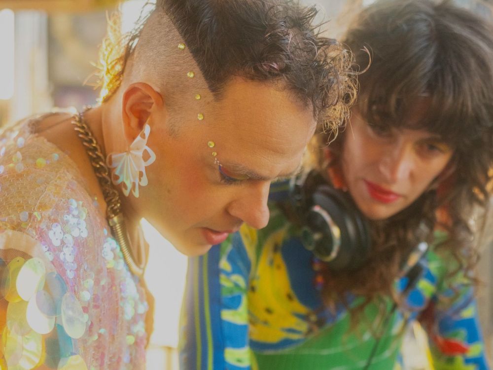 A man and woman in vibrant clothing stand side by side, smiling at the camera in a cheerful outdoor setting.