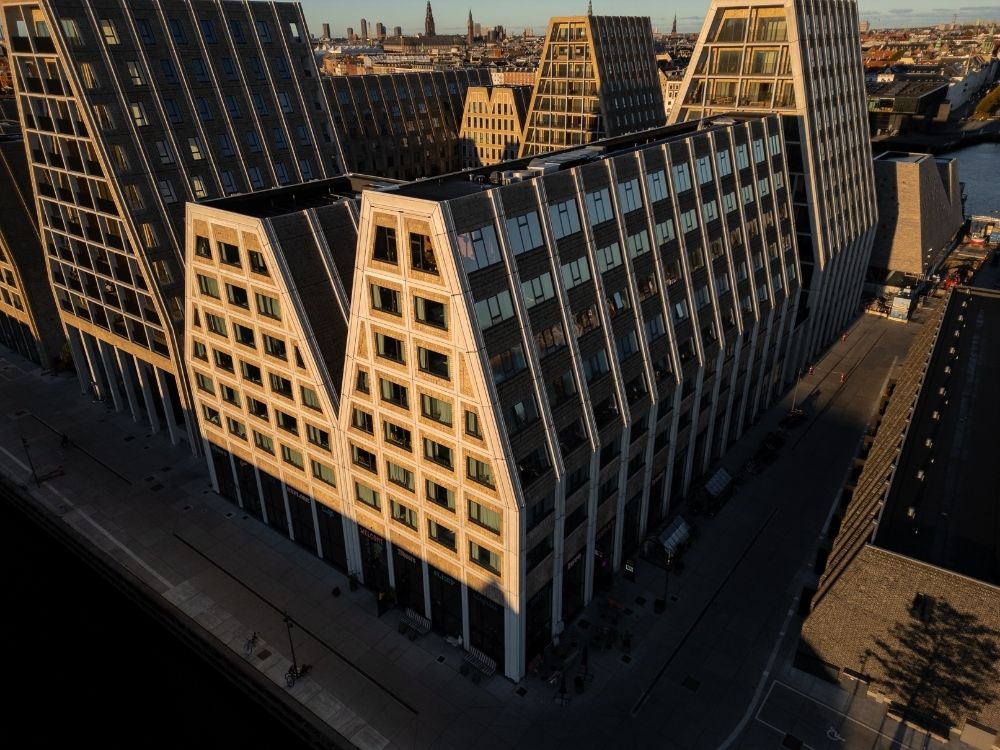 Aerial view of a large, modern building with a distinctive triangular roof pattern at sunset in an urban area.