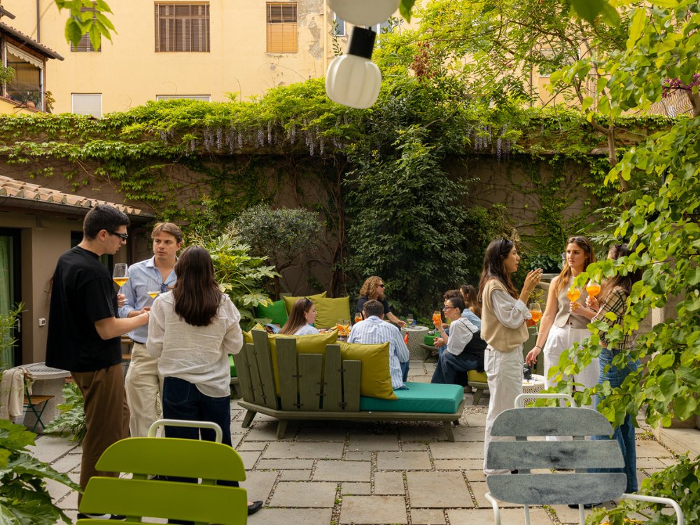Group of people socializing and drinking in a lush, cozy outdoor garden patio with green foliage and comfortable seating.