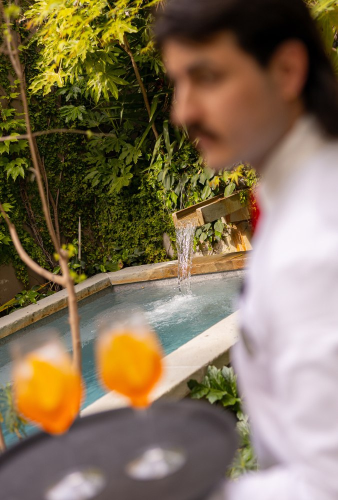 Waiter holding a tray with two orange cocktails near a small outdoor fountain surrounded by green foliage.