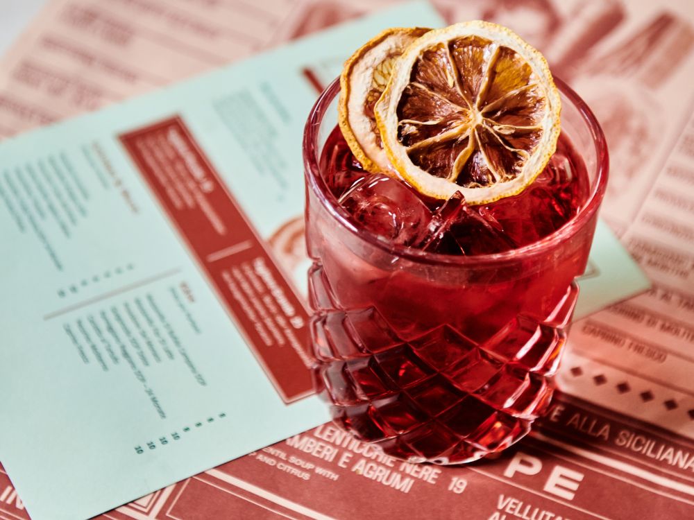Red cocktail in a textured glass with ice and dried citrus slices on top, placed over menus with red and light blue backgrounds.