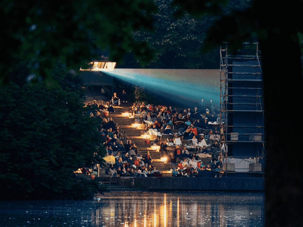 Outdoor cinema event at night with an audience seated on stairs, watching a movie projected across a reflective body of water.