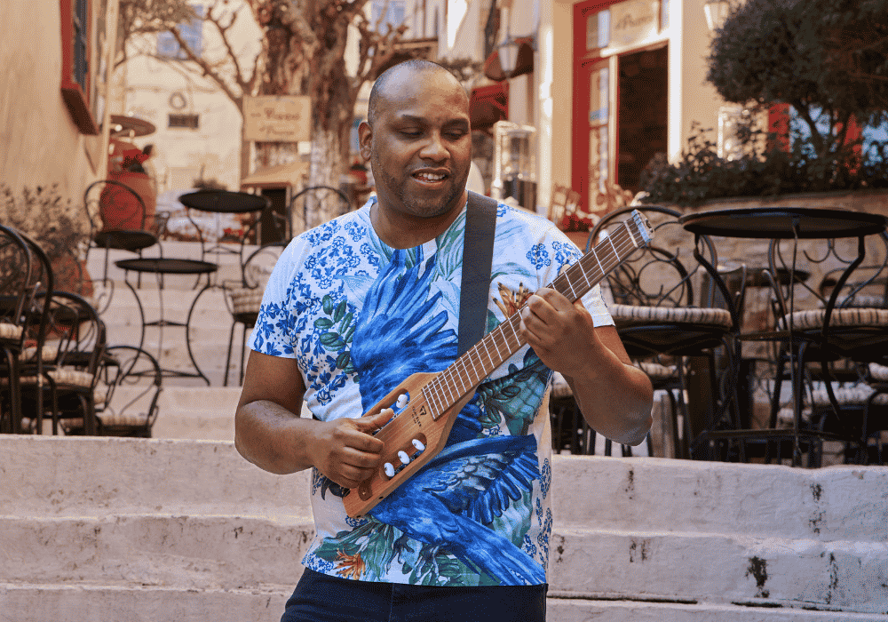Man in a blue patterned shirt playing a ukulele outdoors with cafes and people in the background.