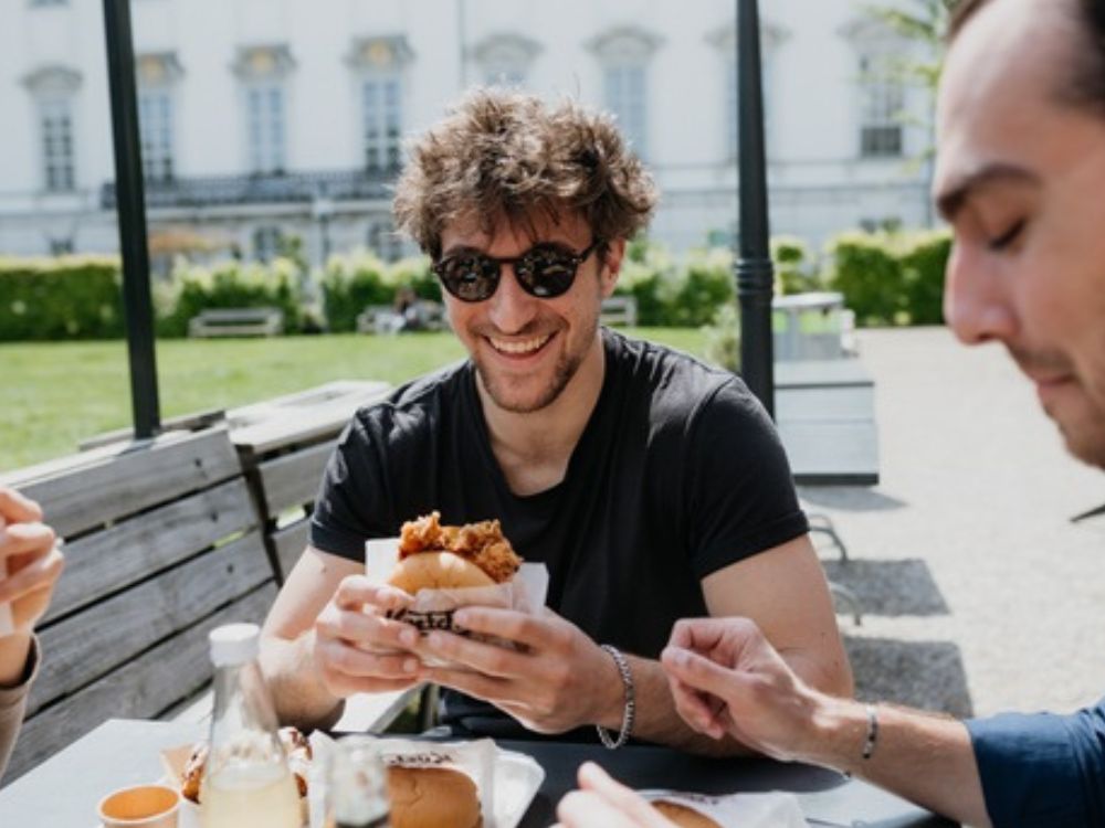 Three individuals enjoying a meal outdoors in a park, surrounded by greenery and a relaxed atmosphere.