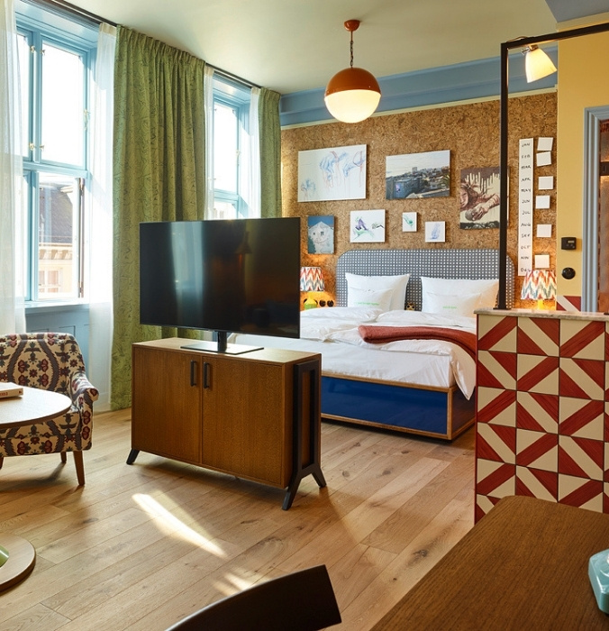 Boutique hotel room featuring a flat-screen TV on a wooden console, a plush king bed with a cork art wall, and a red-and-white patterned room divider.