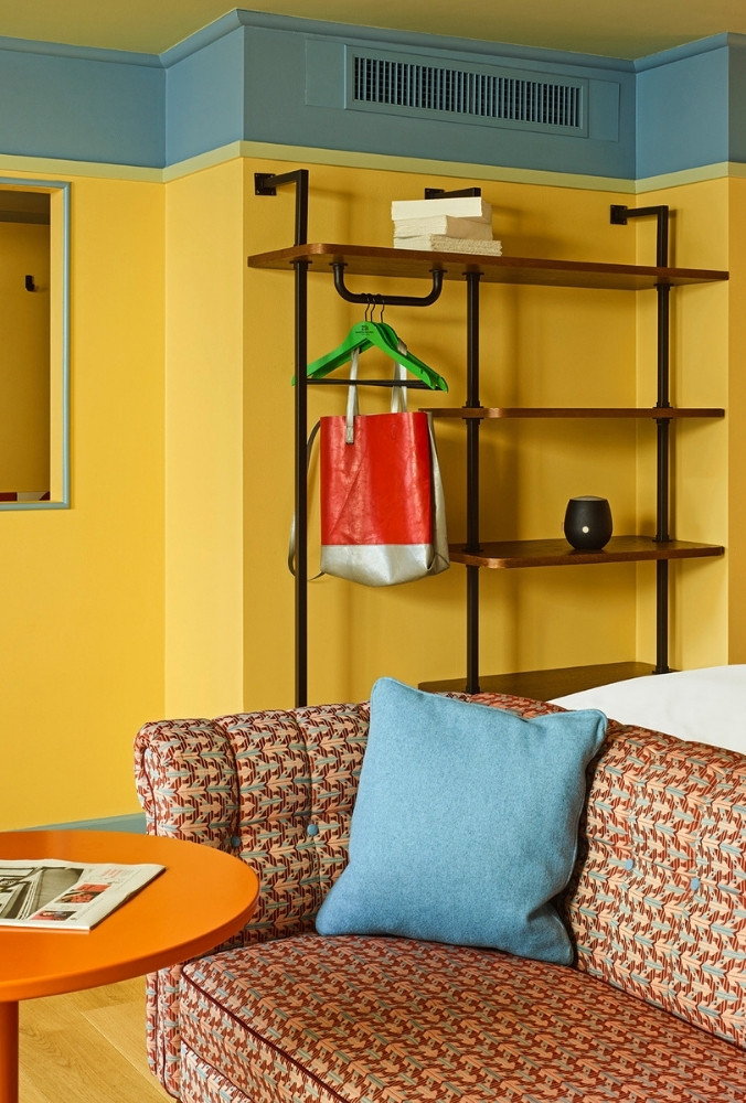 Detail of a patterned fabric sofa with a plush blue cushion and a bright orange coffee table, set against a yellow wall with industrial shelving.