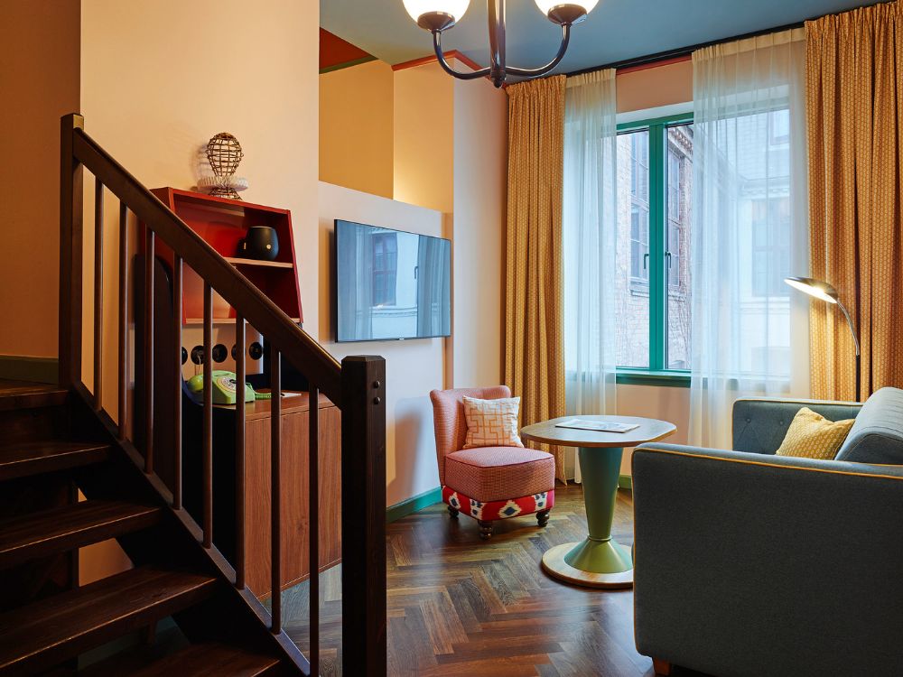 Spacious two-level hotel suite with a dark wood staircase, a wall-mounted TV, a green pedestal table, a blue sofa, and an armchair next to a tall window.