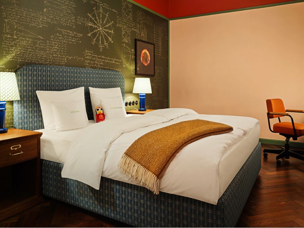 Boutique hotel room featuring twin beds with white duvets, a green chalkboard accent wall with map sketches, and a retro orange office chair.