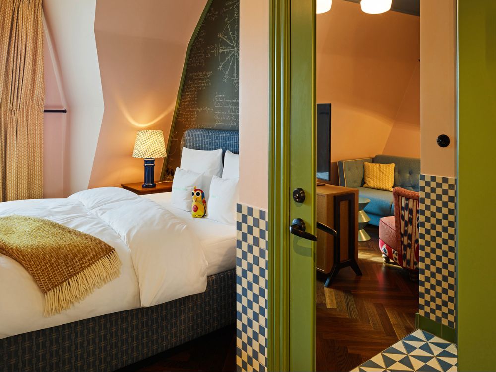 View through a green door frame showing a boutique bedroom on the left and a cozy sitting lounge with a blue sofa and patterned armchair on the right.