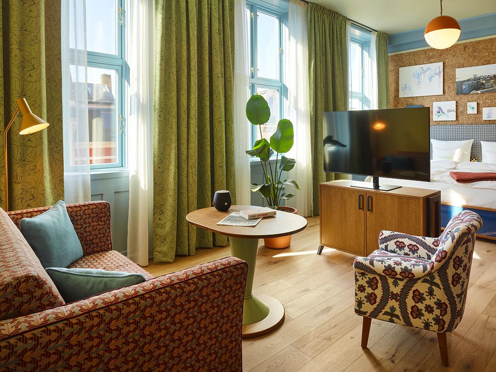 Hotel suite living area showing a flat-screen TV, a patterned fabric sofa, a round green-based table, and a colorful armchair next to tall windows.