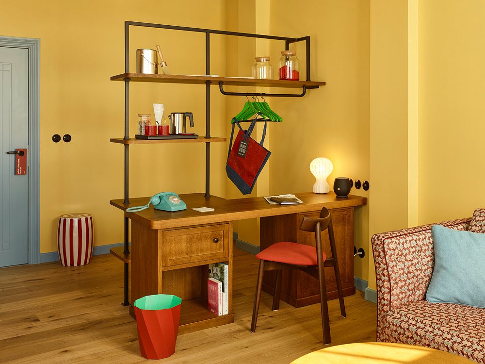 Modern hotel writing desk in a yellow room featuring an integrated industrial shelf, green clothing hangers, and a retro turquoise rotary phone.