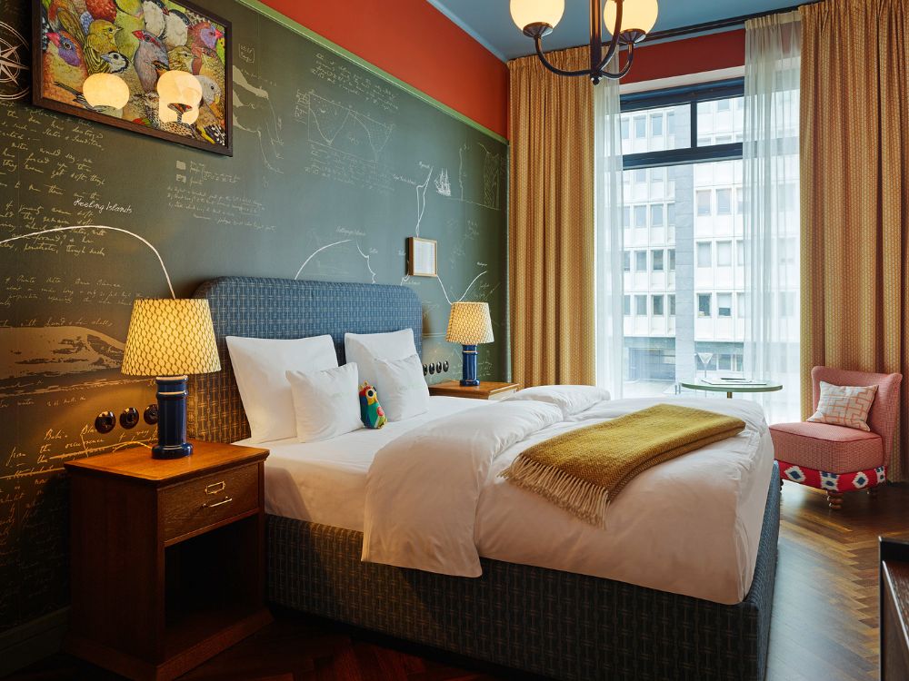Double bed with a blue headboard set against a dark green chalkboard wall decorated with map sketches and classic reading lamps.
