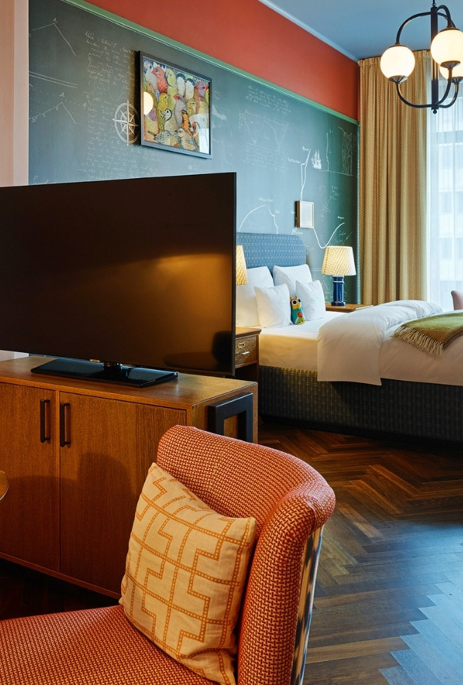 Angle from an orange armchair looking across a television console toward a bed under a dark green chalkboard map wall.