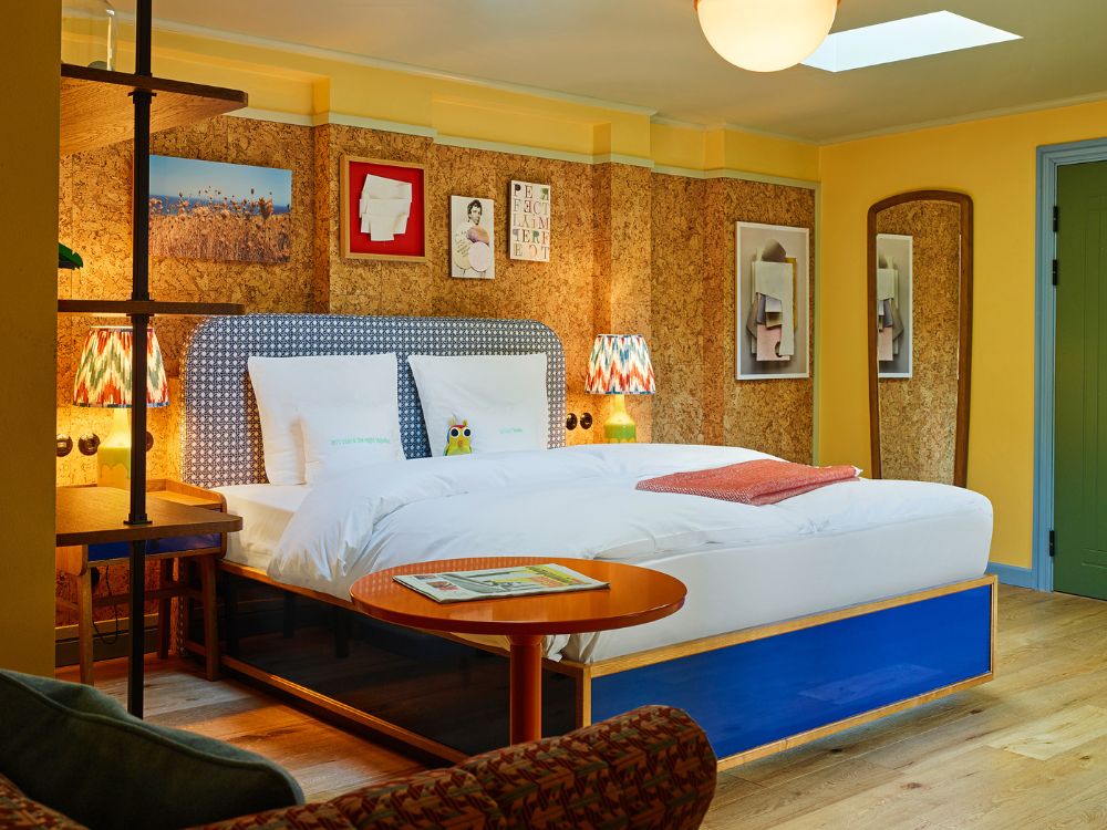 Quirky hotel bedroom showing a king bed set against a cork wall with framed gallery artwork and a sleek orange coffee table in the foreground.