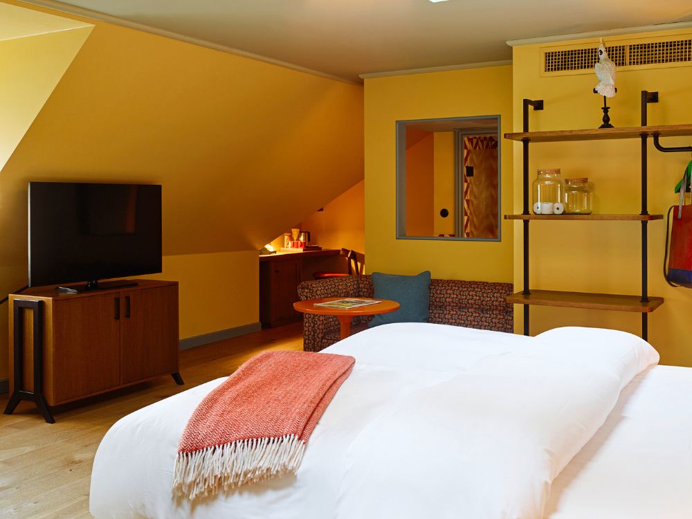 View over a white duvet with a rust-colored blanket looking into a yellow attic suite featuring a flat-screen TV and a cozy sofa seating area.