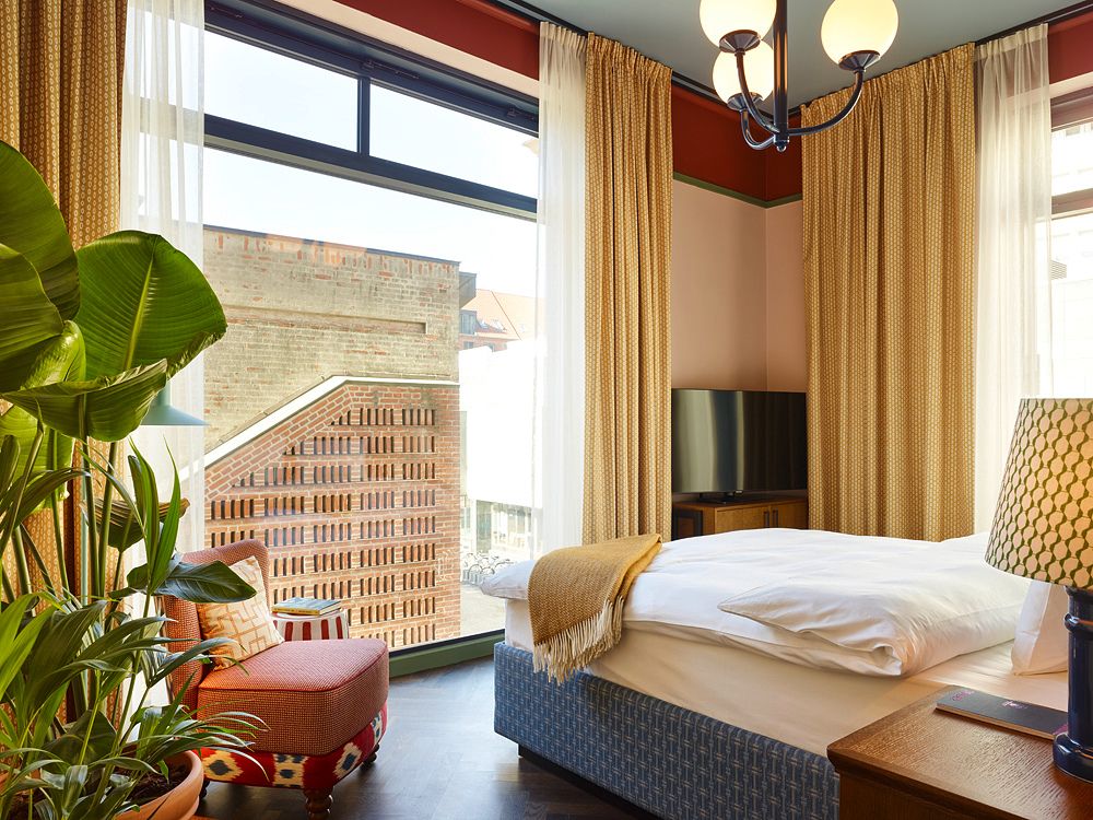 Stylish hotel room featuring a bed with a blue headboard, a large green arched window with yellow curtains, a patterned armchair, and a dark chalkboard wall.