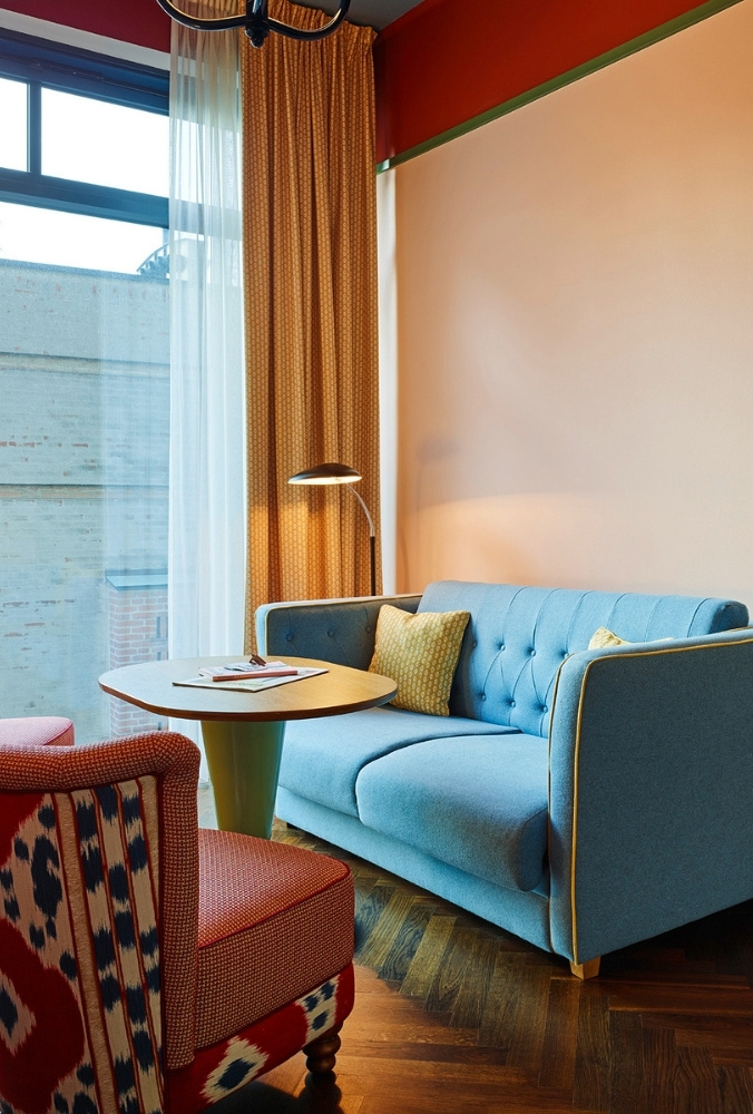 Comfortable hotel suite lounge corner featuring a button-tufted blue fabric sofa, a red patterned armchair, and a retro floor lamp on herringbone parquet floors.