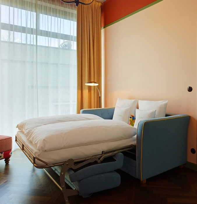 An open-concept hotel room featuring a blue sofa converted into a double bed with white duvets and fluffy pillows next to a sunny window.