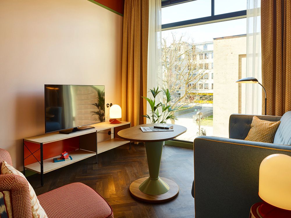Modern guest room seating area with a blue sofa, a round wooden table, a TV on a minimal wooden console, and a large window looking onto a city street.