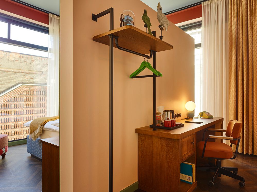 Hotel room study area with a wooden writing desk, an orange swivel chair, a retro table lamp, and an integrated shelf on a room divider wall.