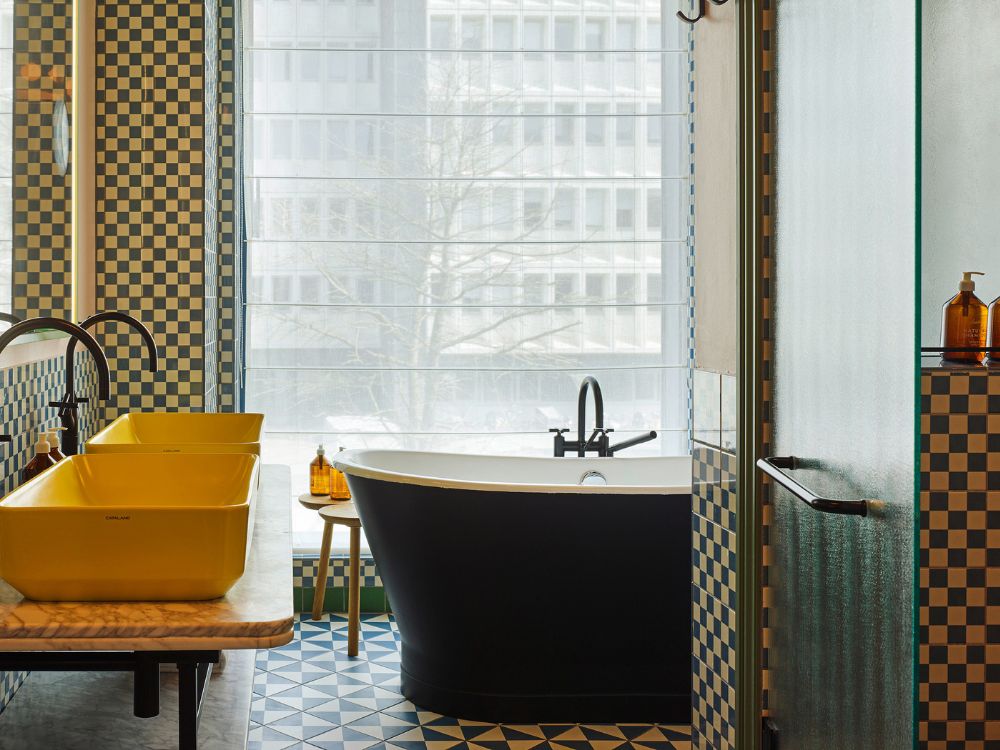 Luxury hotel bathroom featuring a black freestanding bathtub, double yellow vessel sinks on a marble counter, and blue checkered wall tiles next to a window.