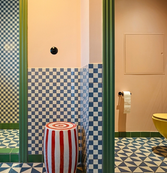 Vibrant hotel bathroom with blue-and-white checkered wall tiles, geometric floor tiles, a yellow toilet, and a red-and-white striped ceramic stool.
