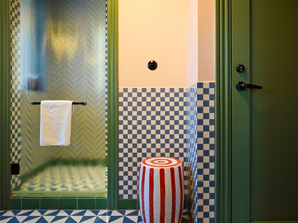 Boutique hotel bathroom featuring a frosted glass shower door with a branded towel, a red-and-white striped ceramic stool, and blue checkerboard tiled walls.