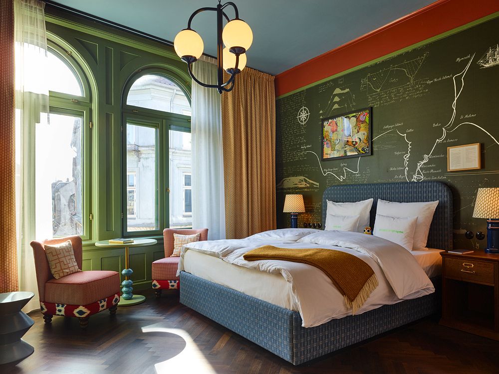 Stunning hotel room interior with high green-painted arched windows, a chalkboard map accent wall, herringbone parquet floors, and cozy pink armchairs.