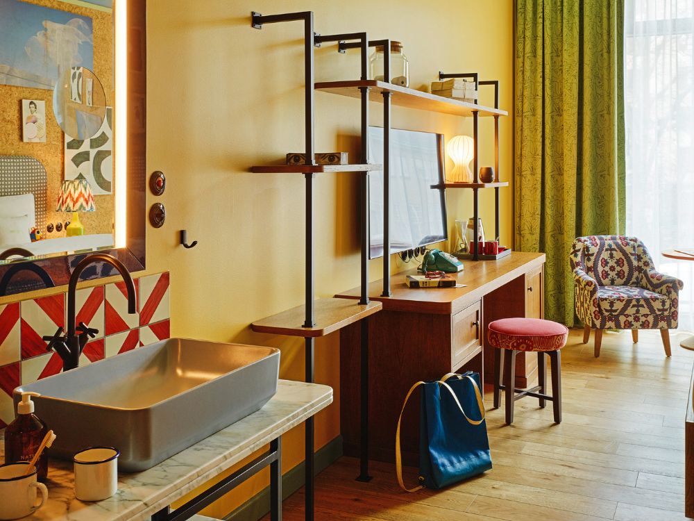 Boutique hotel room interior featuring a marble vanity with a grey vessel sink, a warm yellow wall with industrial shelving over a desk, and a patterned armchair.