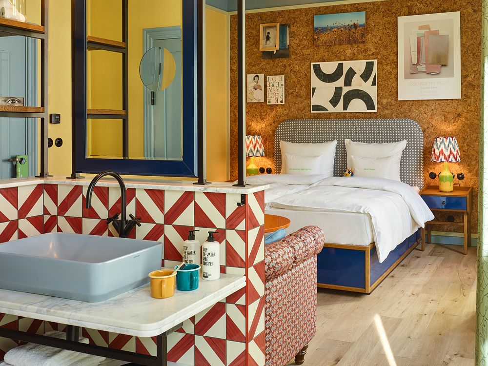 Open-concept guest room vanity with a grey square vessel sink on marble, backed by red and white geometric tiles, looking through to a king bed.