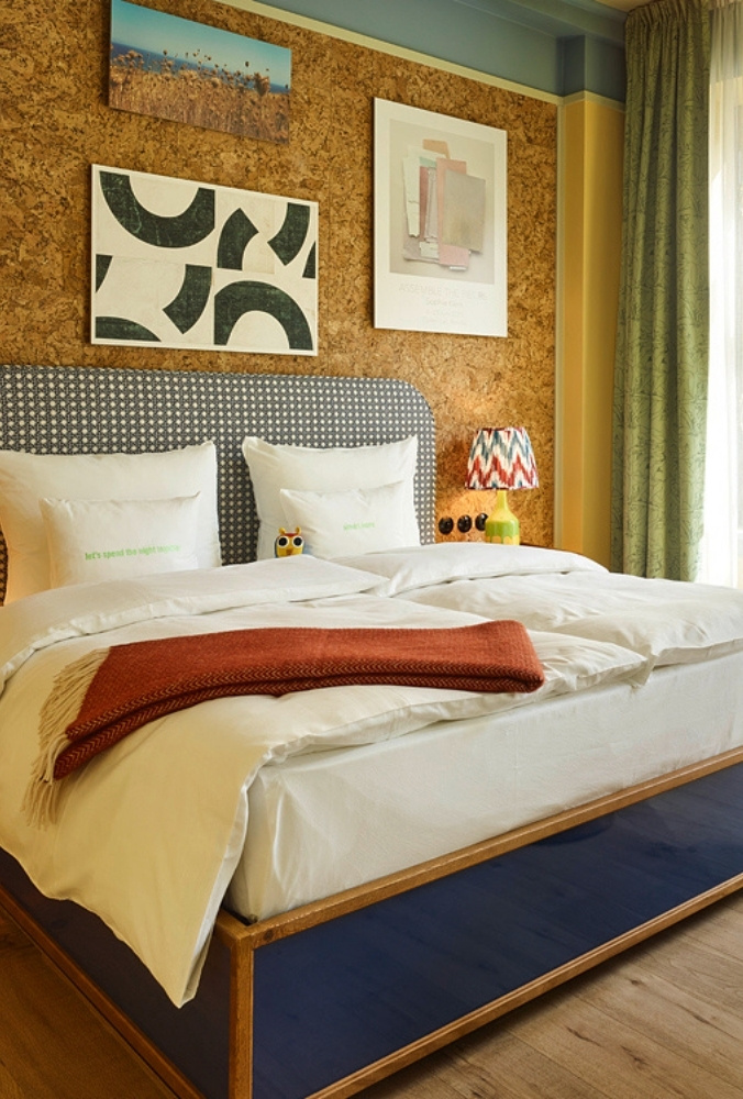 Hotel double bed featuring a blue patterned headboard, white duvet, and a rust-orange blanket, set against a warm cork-paneled wall with framed art.