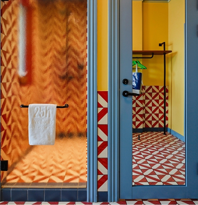 Frosted glass hotel shower door with a branded white towel, reflecting bold red and white geometric wall tiles and a blue door frame.
