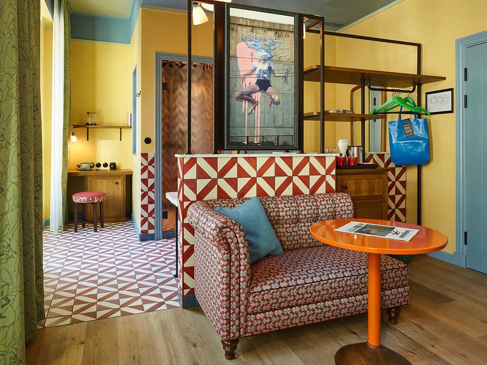 Boutique hotel suite living area featuring a patterned fabric sofa, a round orange wooden table, and a geometric tiled divider wall with art panels.