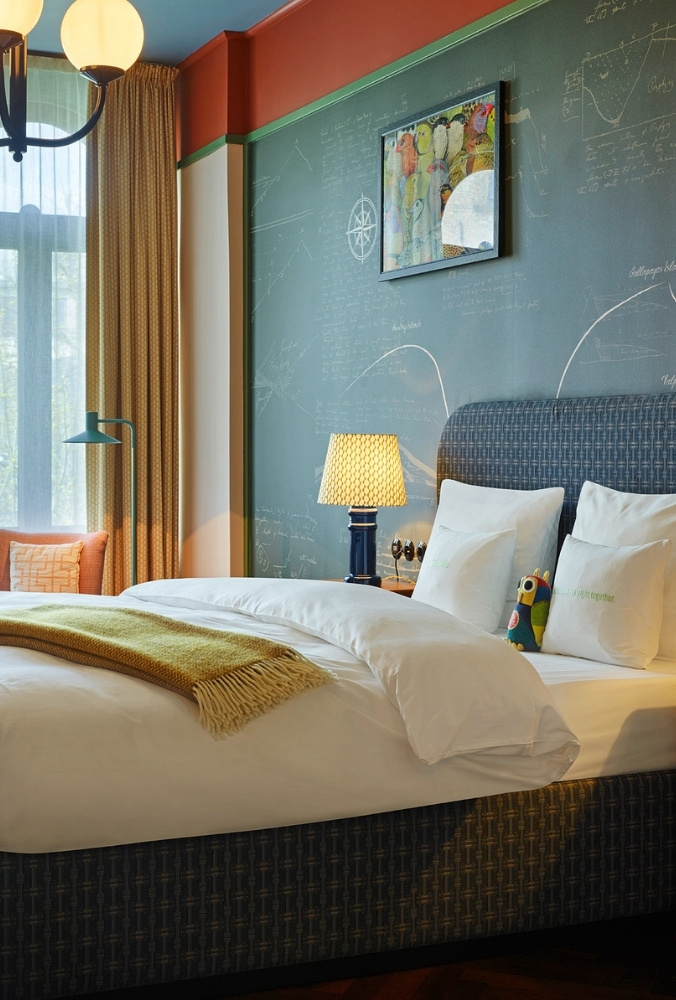 Boutique hotel bed with white linens and a yellow throw blanket, set against a dark green chalkboard wall decorated with chalk illustrations.