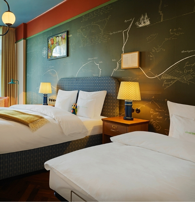 Comfortable hotel twin bedroom featuring a dark green chalkboard wall with map sketches, modern bedside lamps, and a wooden nightstand.