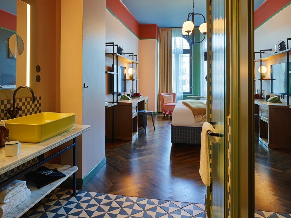 Spacious hotel suite showing a yellow washbasin vanity with blue triangular floor tiles, looking towards dark herringbone wood floors, a desk, and a bed by a tall window.