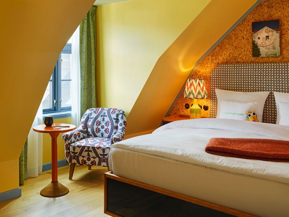 Cosy hotel attic bedroom under a sloped yellow wall, featuring a bed with a cork-patterned headboard, a colorful bedside lamp, and a patterned fabric armchair.