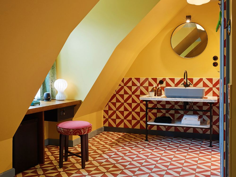 Attic hotel suite workspace and vanity under sloped yellow ceilings, featuring a small desk with a pink stool, a grey vessel sink on marble, and red geometric tiles.