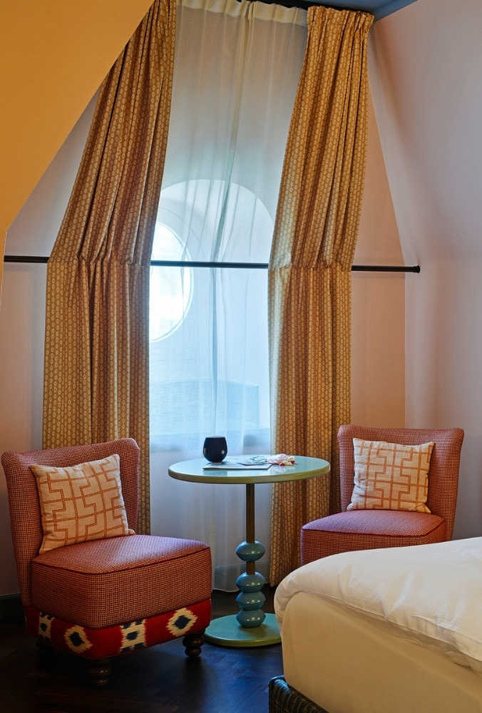Cosy hotel attic sitting nook next to a window featuring two pink upholstered armchairs, patterned pillows, and a teal pedestal table.