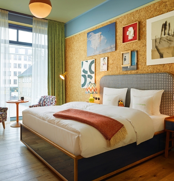Bright hotel room featuring a king bed with a patterned headboard, a golden-brown throw blanket, and a warm cork-paneled accent wall with framed artwork.