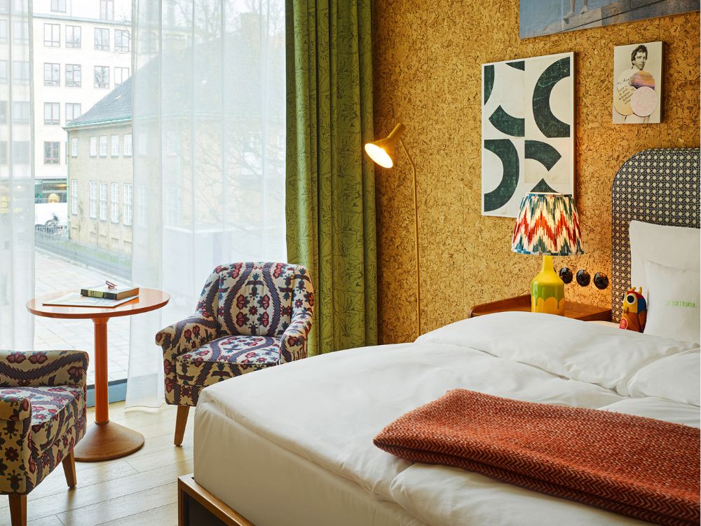 Cosy hotel bedroom corner by a large window featuring two patterned fabric armchairs, a small orange table, and a bed with a cork wall behind it.