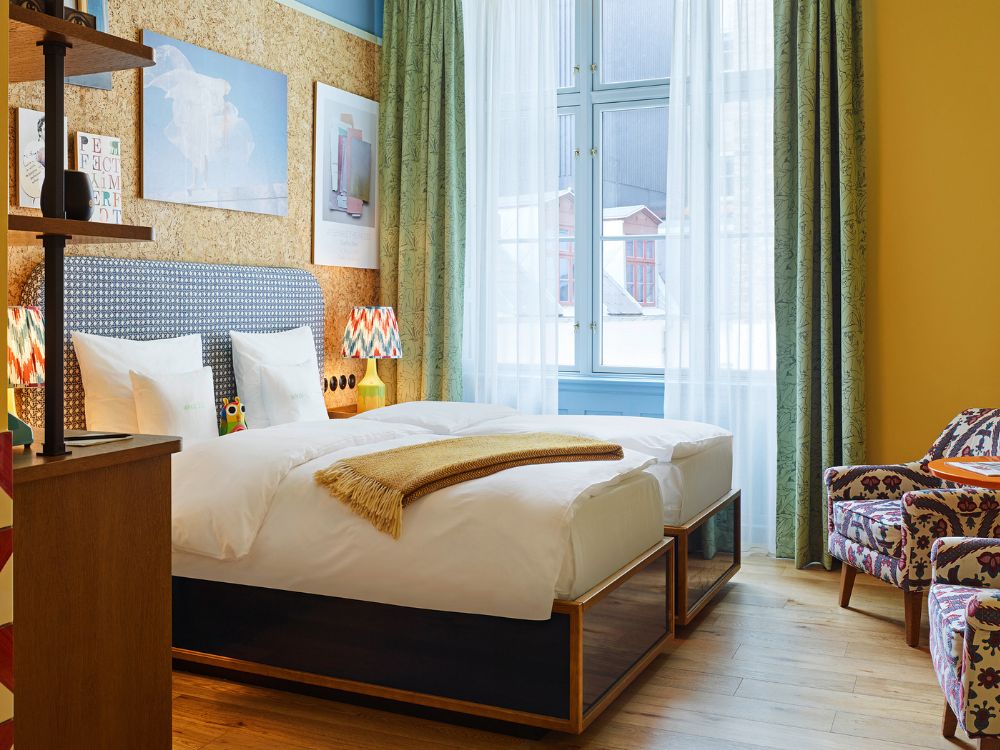 Bright hotel room with two twin beds next to a large window, featuring green patterned curtains, a cork wall with artwork, and a colorful upholstered armchair.